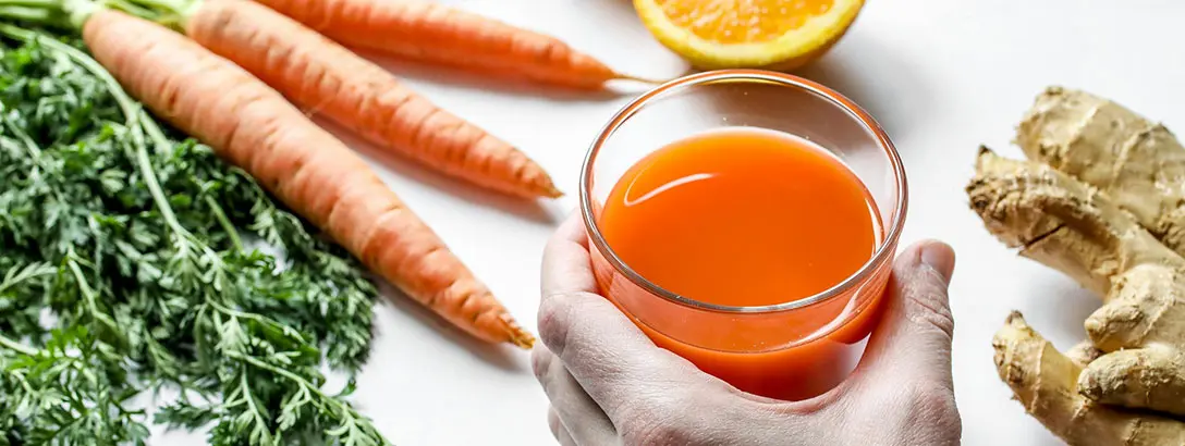 A glass of carrot with ginger juice surrounded by carrot and ginger