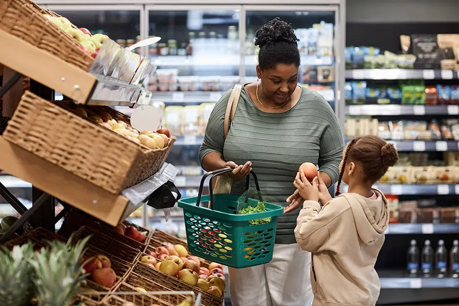A woman and a child are shopping in a grocery store, surrounded by wooden baskets of apples and other produce. The woman holds a green shopping basket while the child offers her an apple, conveying a warm and engaging shopping moment.