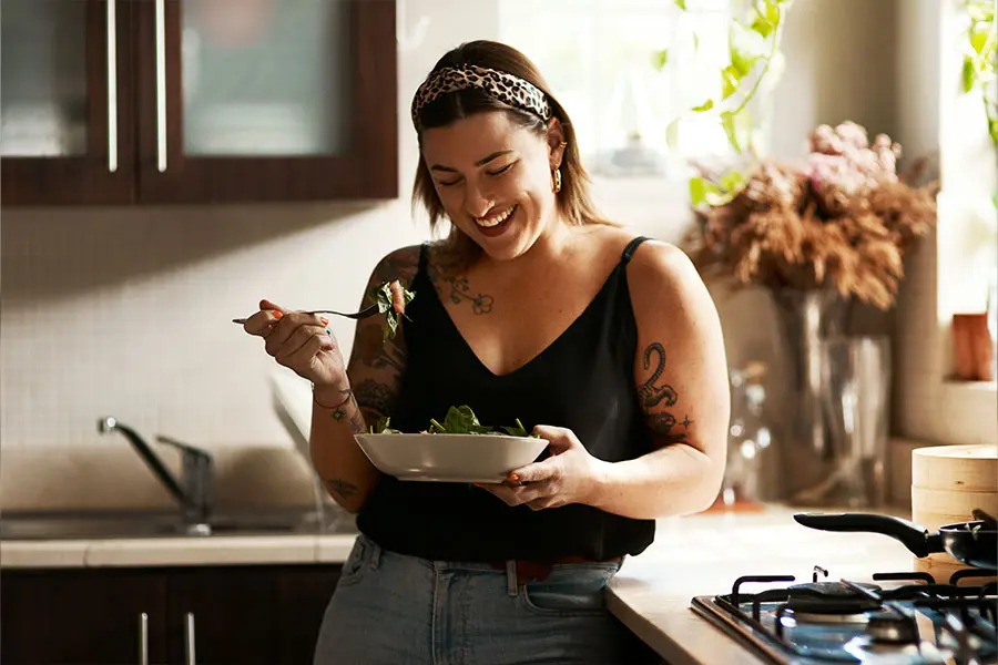 A woman with tattoos smiles as she enjoys a salad in a bright kitchen. She holds a fork with greens in one hand and a bowl of salad in the other, radiating joy.