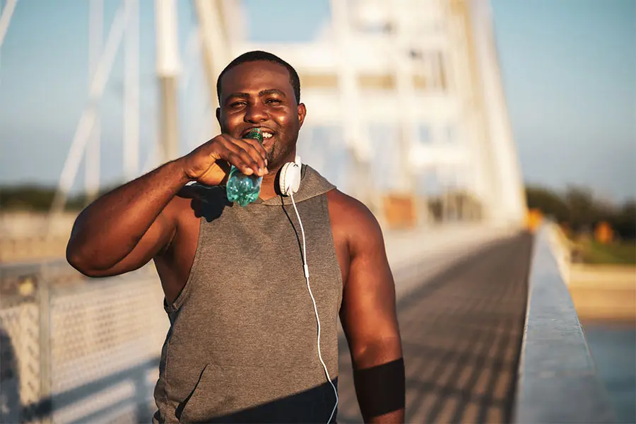 A man in a grey sleeveless hoodie stands on a bridge, smiling while drinking from a water bottle. He wears white headphones and is bathed in warm sunlight, suggesting a relaxed and active mood.