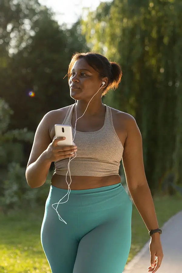 A woman walks along a sunlit path wearing a grey crop top and turquoise leggings, holding a smartphone with earphones. Lush greenery surrounds her, suggesting an active and relaxed mood.