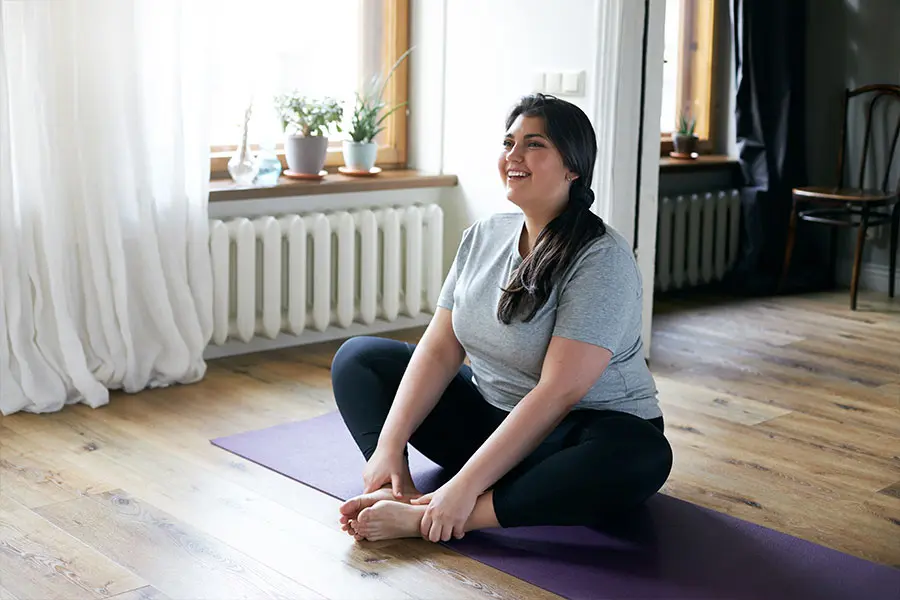 A person with long dark hair sits on a purple yoga mat in a bright, airy room, smiling while stretching their feet. Sunlight streams through the large windows, illuminating greenery on the windowsill.