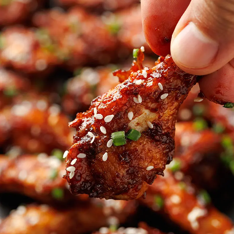 A close-up of a hand holding a sticky, glistening chicken wing covered in a spicy glaze, topped with sesame seeds and green onions, with other wings blurred in the background.