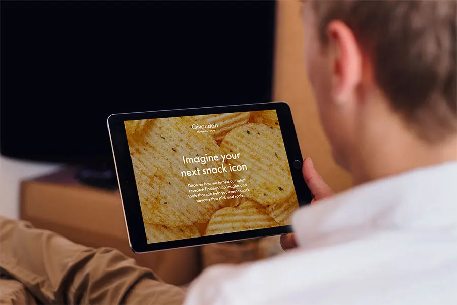 A person holds a tablet displaying a website from Givaudan with the text "Imagine your next snack icon" over a background of potato crisps. The setting is relaxed, with a blurred background suggesting a home environment
