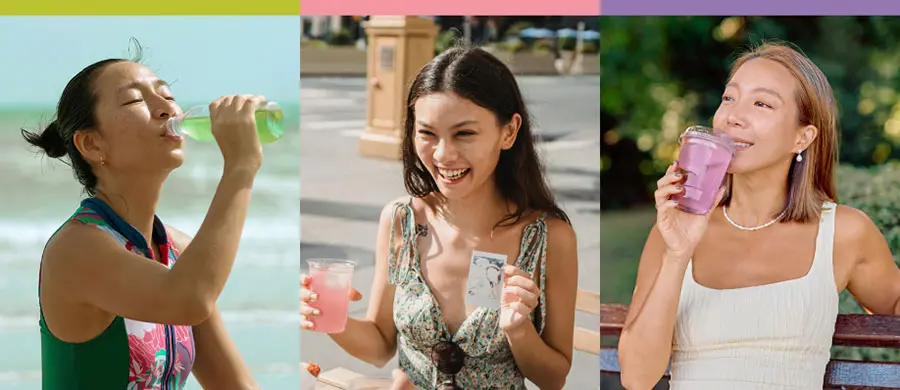 Three women enjoy refreshing drinks in a vibrant outdoor setting. The first woman, wearing a colourful swimsuit, sips a green drink by the beach. The second, in a floral dress, smiles while holding a transparent cup. The third woman, seated on a bench, enjoys a pink beverage, conveying a joyful, relaxed atmosphere.