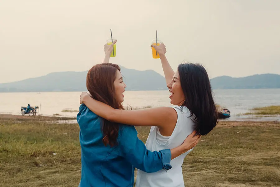 Two women are joyfully celebrating by a riverbank, holding up drinks in vibrant yellow and green cups. They are embracing and laughing, with mountains in the background and a warm, hazy sky.