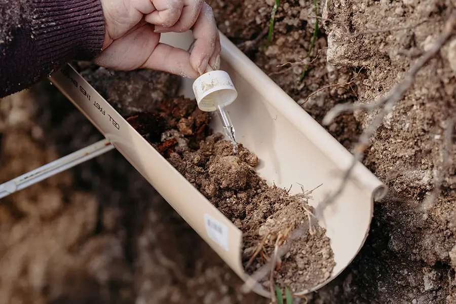 A hand holds a dropper above a trough filled with soil, used for testing. The background shows earthy tones and green grass, suggesting an agricultural setting.