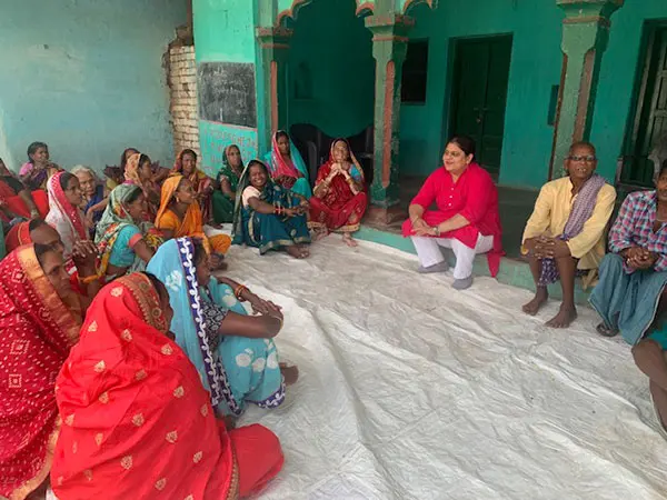 A group of women in vibrant traditional clothing sits on a white cloth, engaging in discussion. In the foreground, a woman in a pink top listens attentively, while others appear animated and involved. The setting features turquoise walls and an open doorway, creating a warm and inviting atmosphere.
