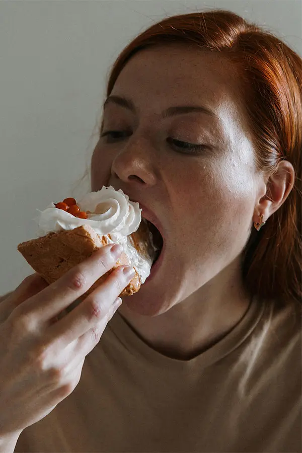 A woman with red hair is seen about to take a big bite of a dessert topped with whipped cream and red berries. She is wearing a beige top and has a joyful expression, emphasizing the enjoyment of the treat.