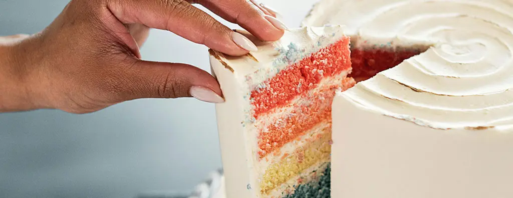 A hand is lifting a slice from a visually striking layered cake, showcasing vibrant layers of red, orange, yellow, and blue cake beneath a smooth white icing. The setting has a soft background, evoking a cheerful and inviting mood.