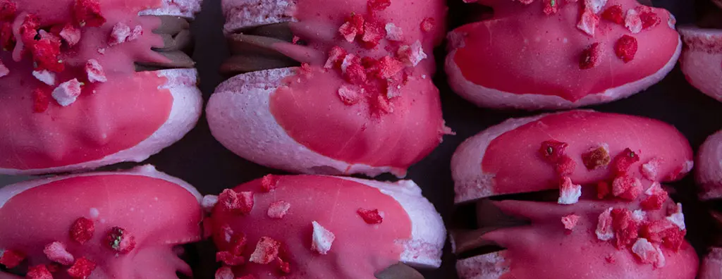 Delicious pink desserts with a glossy chocolate filling, topped with crushed freeze-dried strawberries. The sweets are arranged neatly against a dark background, enhancing their vibrant colours.