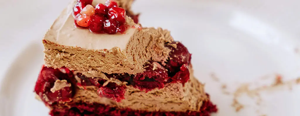 A slice of rich chocolate mousse cake sits on a white plate, topped with glossy red berries. The layered texture reveals a chocolate filling interspersed with fruit, evoking a feeling of indulgence.