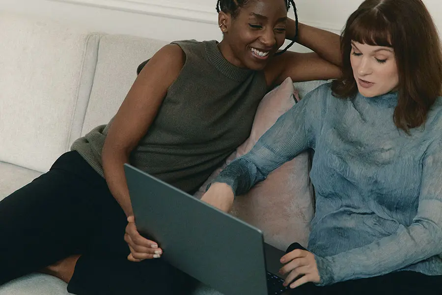 Two women sit together on a light-coloured sofa, smiling as they look at a laptop screen. One wears a dark sleeveless top, while the other is in a blue long-sleeved top. The mood is relaxed and collaborative.