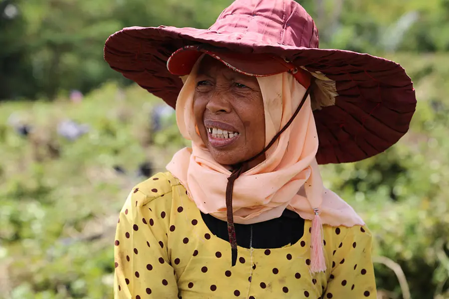 A woman in a large red sun hat and a yellow polka dot blouse sits in a lush green field, showing a thoughtful expression. She wears a light-coloured scarf around her neck, with patches of greenery and blurred figures working in the background.