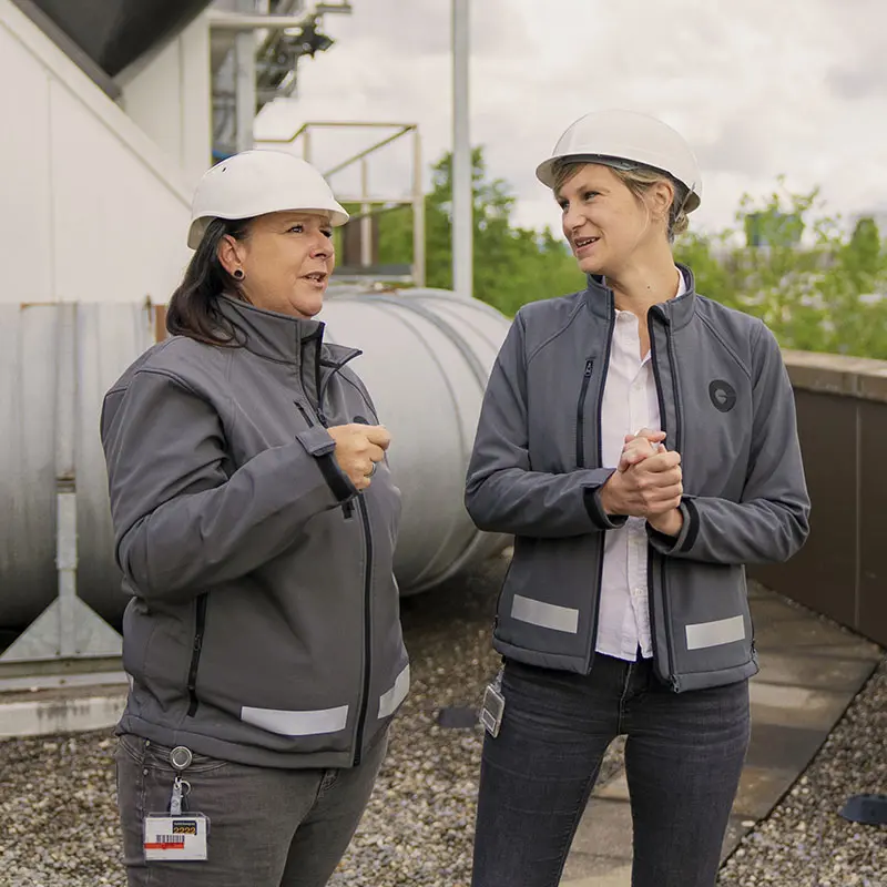 Two women wearing grey jackets and white hard hats converse on a rooftop. They are surrounded by industrial equipment and greenery, indicating a collaboration on operations.