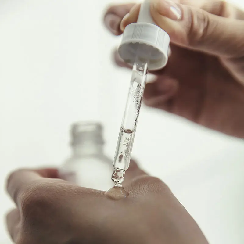 A hand holds a dropper above a wrist, releasing a clear serum onto the skin. The background is softly blurred, creating a clean and minimalist aesthetic.