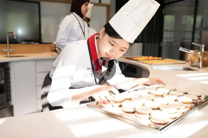 A young chef wearing a white uniform and tall hat carefully arranges dessert sandwiches on a tray in a modern kitchen. Another chef is seen in the background, suggesting a collaborative atmosphere.