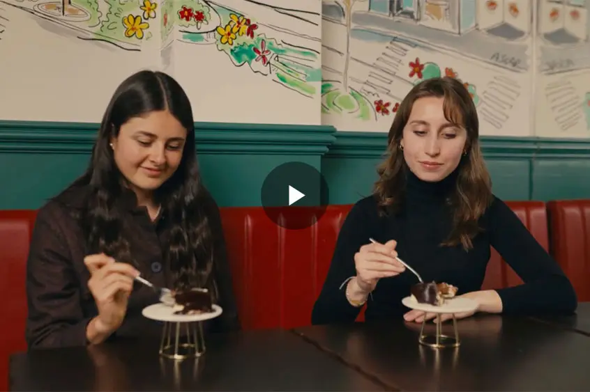 Two women sit at a table in a café, enjoying chocolate desserts on white plates. The setting features colourful artwork on the walls and a maroon banquette. Both appear relaxed and happy.