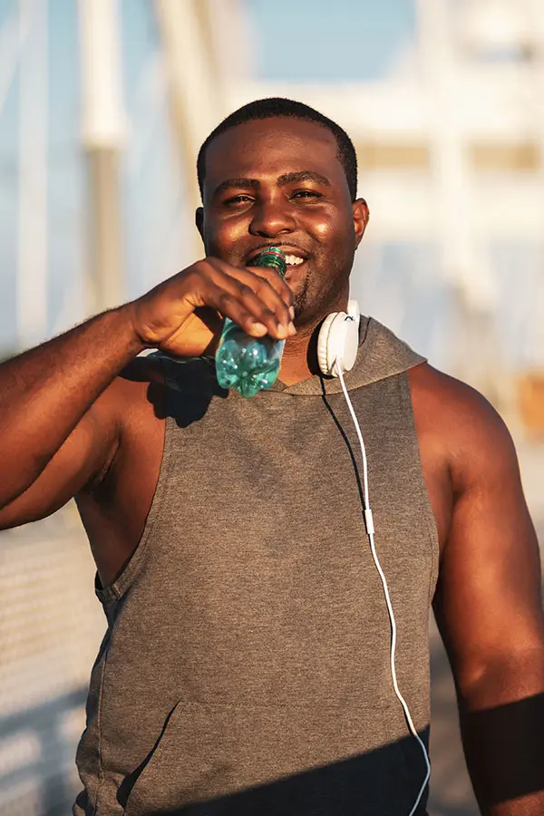 A smiling man in a grey sleeveless hoodie drinks from a green water bottle while wearing white headphones. The bright outdoor setting suggests an active, healthy lifestyle.