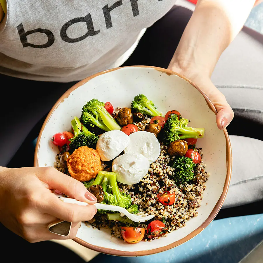 A person seated on a blue mat holds a bowl filled with quinoa, cherry tomatoes, broccoli, poached eggs, and a dollop of orange sauce. 