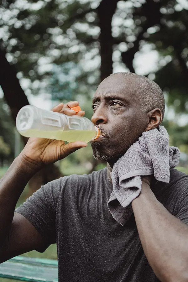A man drinks from a clear sports bottle while wiping his neck with a towel, set against a blurred background of trees. His focused expression reflects a moment of hydration after exercise.