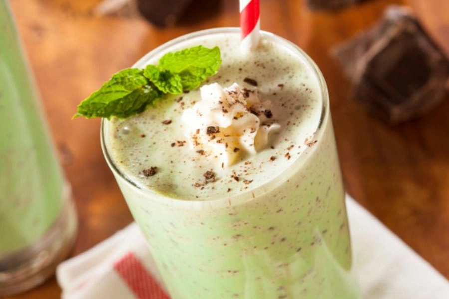 A creamy green mint coloured shake topped with whipped cream, chocolate shavings, and fresh mint, served in a tall glass with a striped straw.