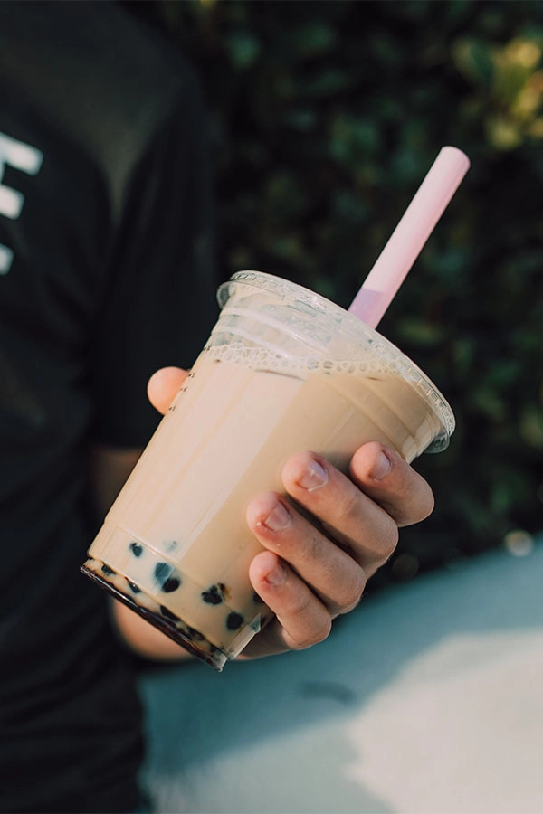 A person in a black “Explore” T-shirt holds creamy iced bubble tea with tapioca pearls and a pink straw, against leafy greenery.