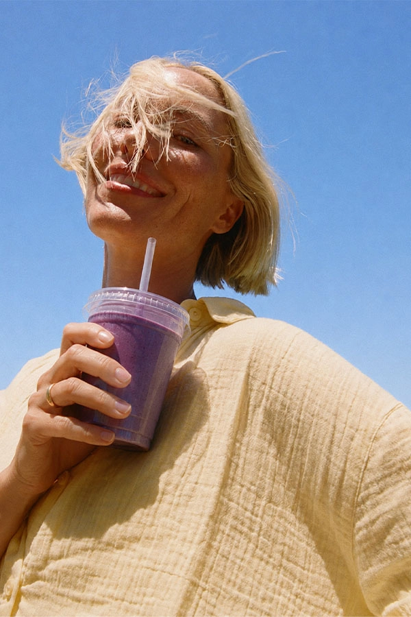 A smiling woman in a textured cream shirt holds a purple smoothie with a straw under a clear blue sky; windblown hair adds a carefree, sunny mood.