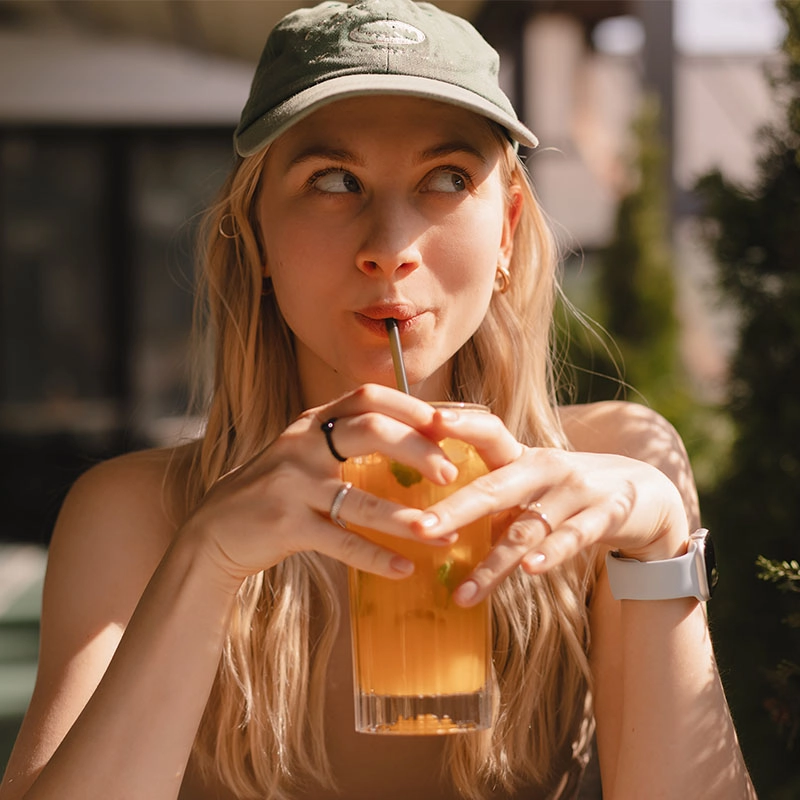 A young woman in a green cap sips an orange drink through a straw, glancing playfully aside in warm sunlight at an outdoor café.
