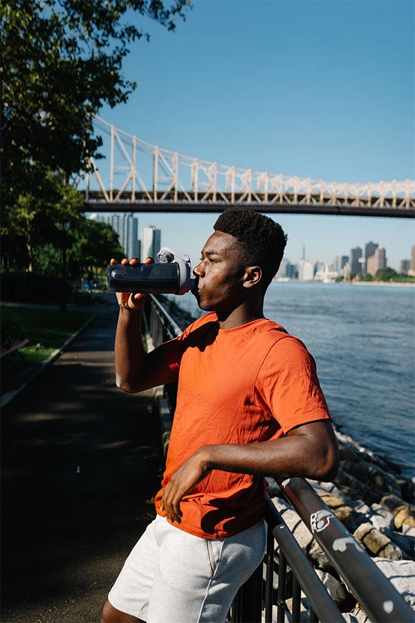 A focused runner in an orange shirt drinks from a water bottle beside a riverside path, with a bridge and city skyline in the sunny background.