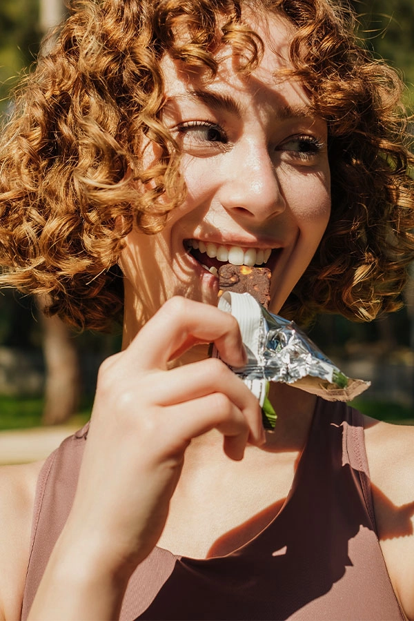 A young curly-haired woman beams as she bites a chocolate bar in silver foil, enjoying a carefree sunny moment among green trees.