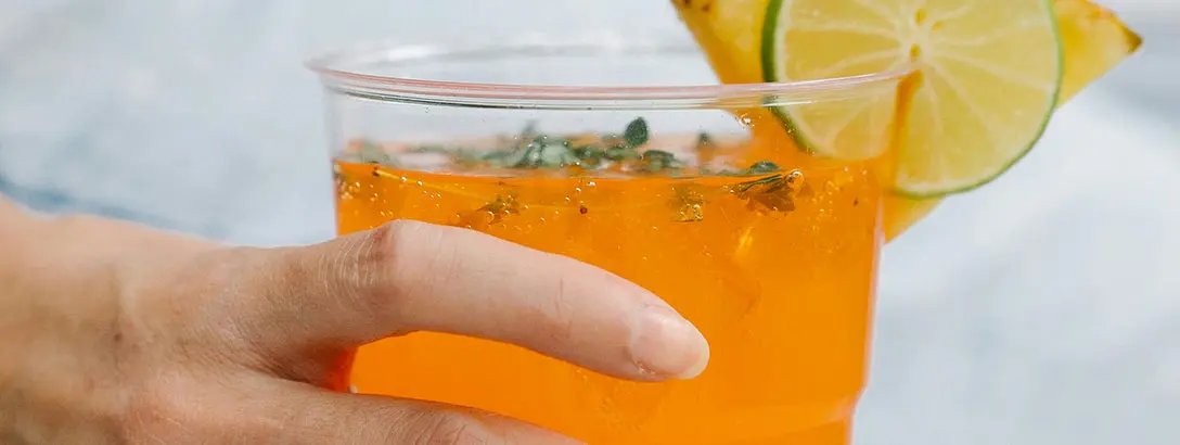 A person holding an orange beverage with a slice of limon and herbs