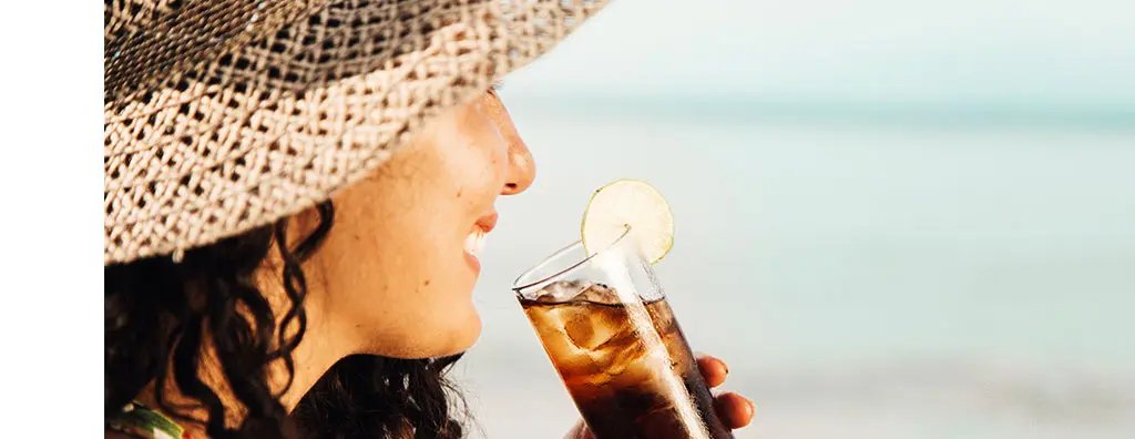 Young woman with sun hat sitting at the beach with a glass of cola with a slice of lemon