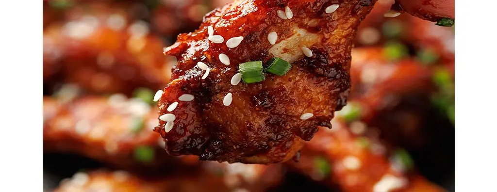 A close-up of a hand holding a sticky, glistening chicken wing covered in a spicy glaze, topped with sesame seeds and green onions, with other wings blurred in the background.