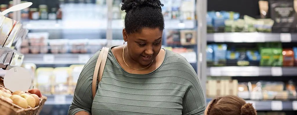 A woman and a child are shopping in a grocery store, surrounded by wooden baskets of apples and other produce. The woman holds a green shopping basket while the child offers her an apple, conveying a warm and engaging shopping moment.