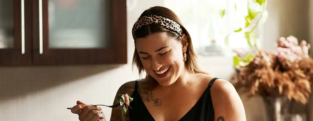 A woman with tattoos smiles as she enjoys a salad in a bright kitchen. She holds a fork with greens in one hand and a bowl of salad in the other, radiating joy.