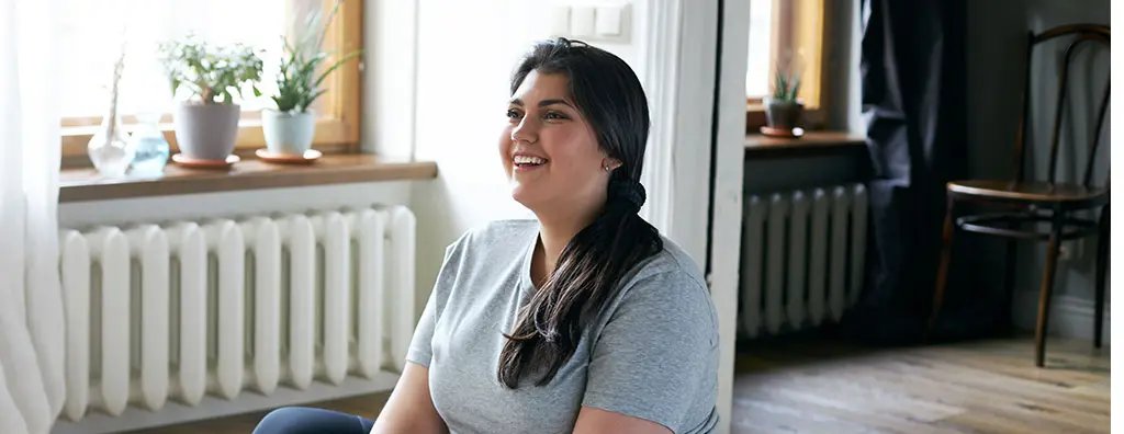 A person with long dark hair sits on a purple yoga mat in a bright, airy room, smiling while stretching their feet. Sunlight streams through the large windows, illuminating greenery on the windowsill.