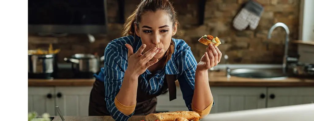 A woman in a blue striped shirt savours a bite of food, with a playful expression, while standing in a warm kitchen filled with cooking pots. A loaf of bread is visible on the countertop.