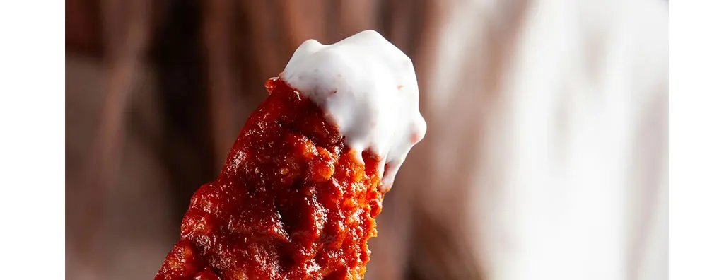 A close-up of a spicy chicken wing coated in bright red sauce, topped with a dollop of creamy white dip. The blurred background suggests a casual dining setting, evoking a tempting and indulgent mood.