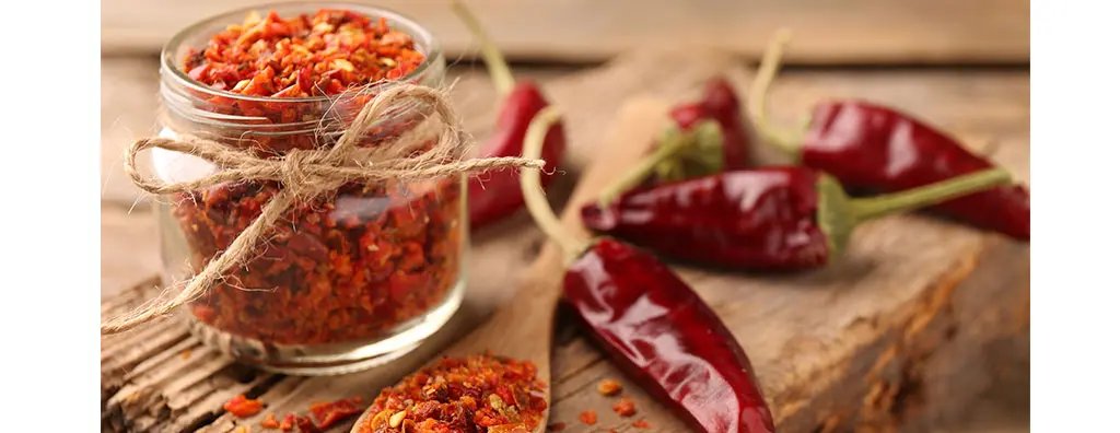 A glass jar filled with vibrant red chilli flakes is tied with twine, surrounded by whole red chillies on a rustic wooden surface. The warm colours and natural setting suggest a focus on spicy flavours.