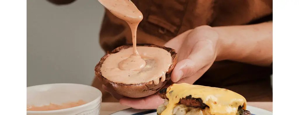 A person is drizzling a spicy sauce onto a toasted brown bun in a kitchen setting. The bun is held in one hand, while a burger with melted cheese sits nearby on a plate. A small bowl of sauce is also visible.