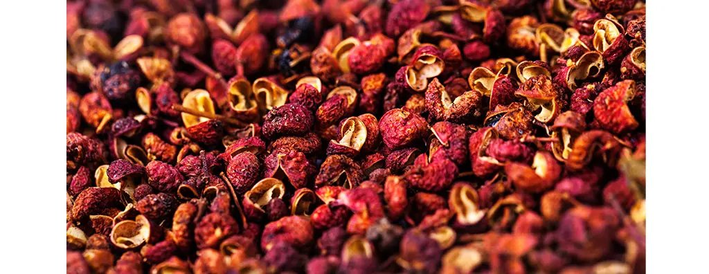 A close-up image of dried Szechuan peppercorns, showcasing their vibrant reddish-brown hues and textured surfaces. The cluster highlights the unique, bumpy shapes of the peppercorns, suggesting a bold, spicy flavour.