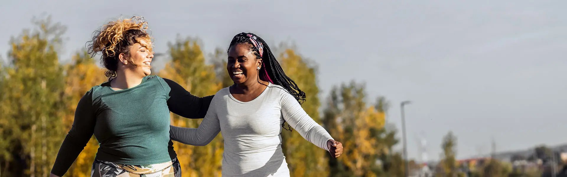 Two women happily jog together outdoors, surrounded by trees with autumn foliage. One wears a long-sleeved green top while the other is in a white top, both smiling joyfully.