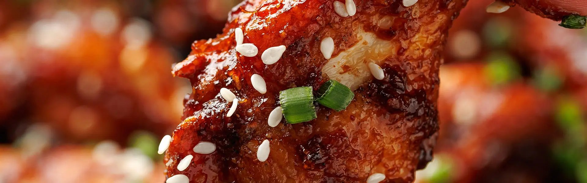 A close-up of a hand holding a sticky, glistening chicken wing covered in a spicy glaze, topped with sesame seeds and green onions, with other wings blurred in the background.
