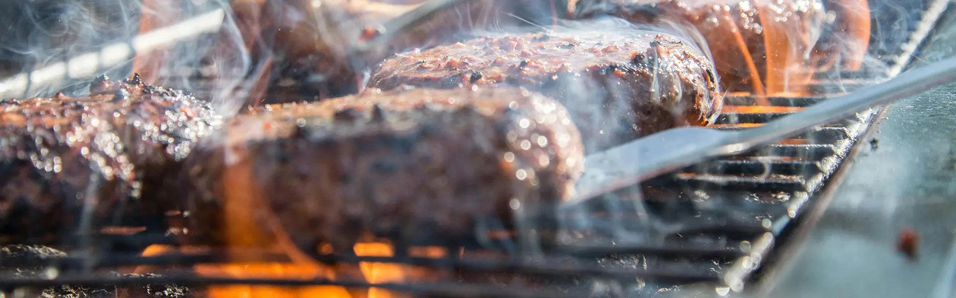 Sizzling burgers cook on a grill, surrounded by swirling smoke and a bright outdoor backdrop. A pair of tongs flips the patties, highlighting a summer barbecue atmosphere.