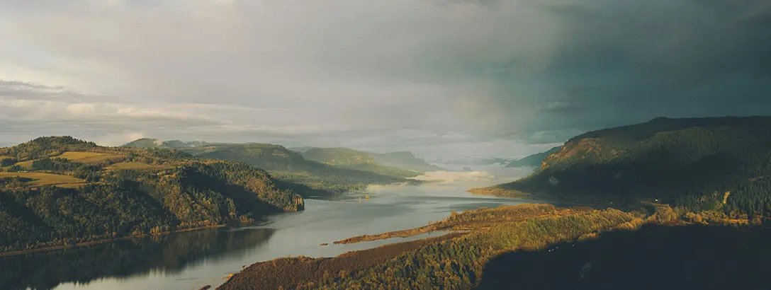 A serene landscape featuring a winding river surrounded by lush green hills under a cloudy sky. Gentle light breaks through the clouds, casting a calm mood.