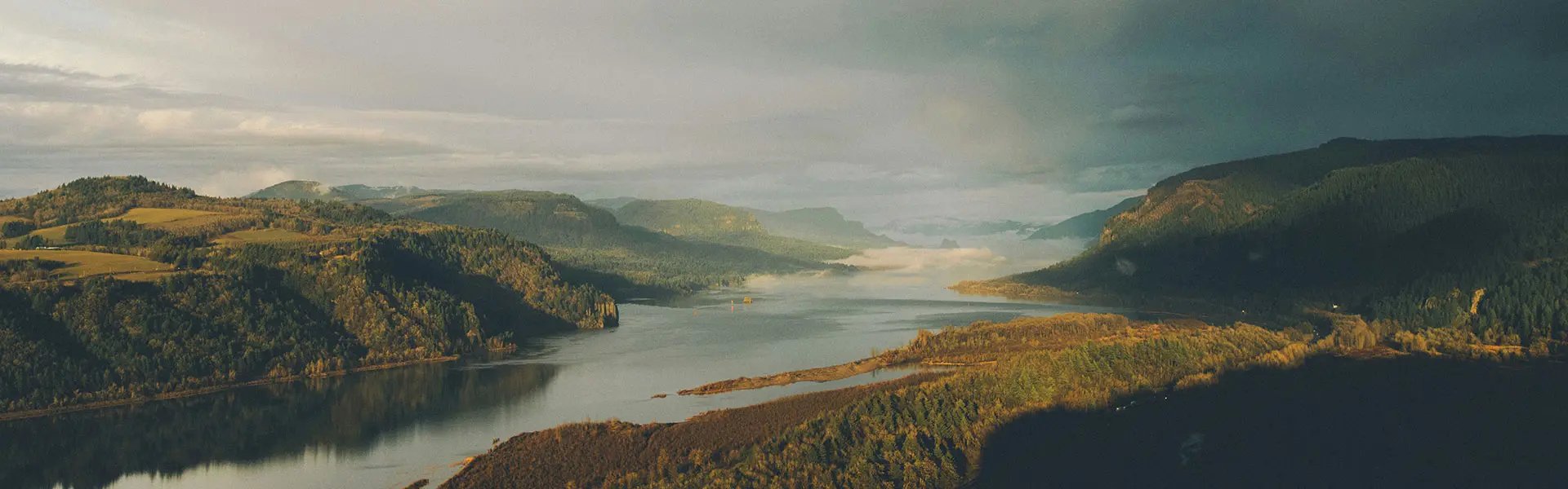 A tranquil landscape featuring a winding river surrounded by lush green hills, under a sky with blue and grey clouds suggesting an impending storm. The scene evokes a sense of calm and anticipation.
