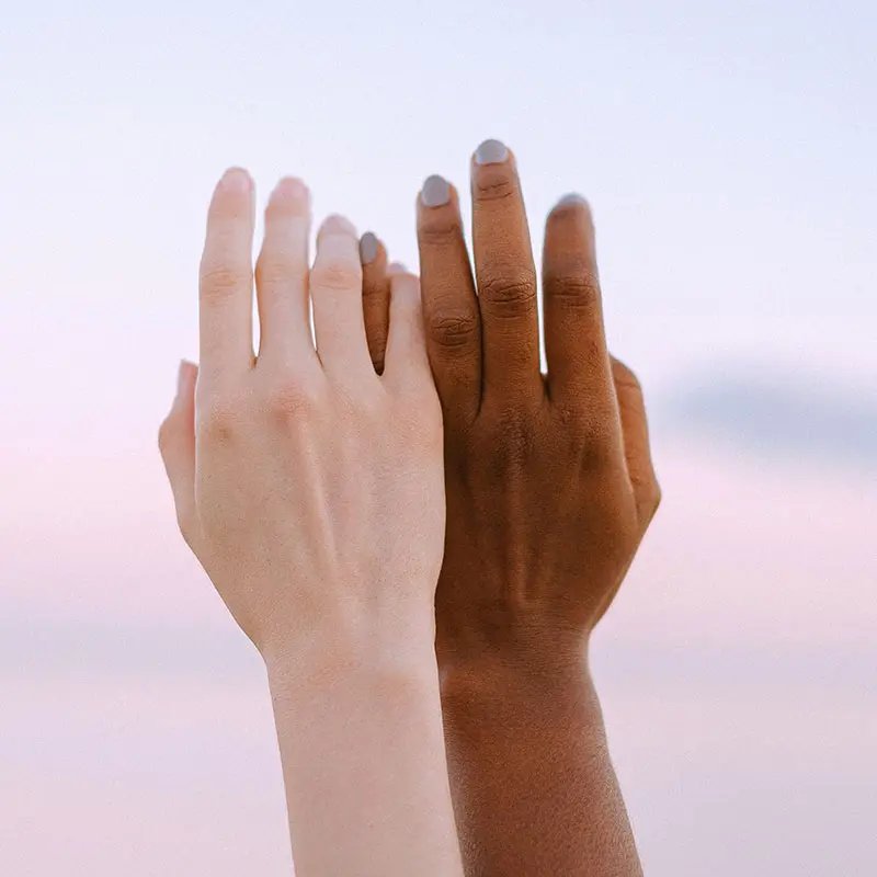 Two hands are raised together against a soft pastel sky, one hand with fair skin and polished nails, the other with dark skin and a natural look. The mood is warm and inclusive.
