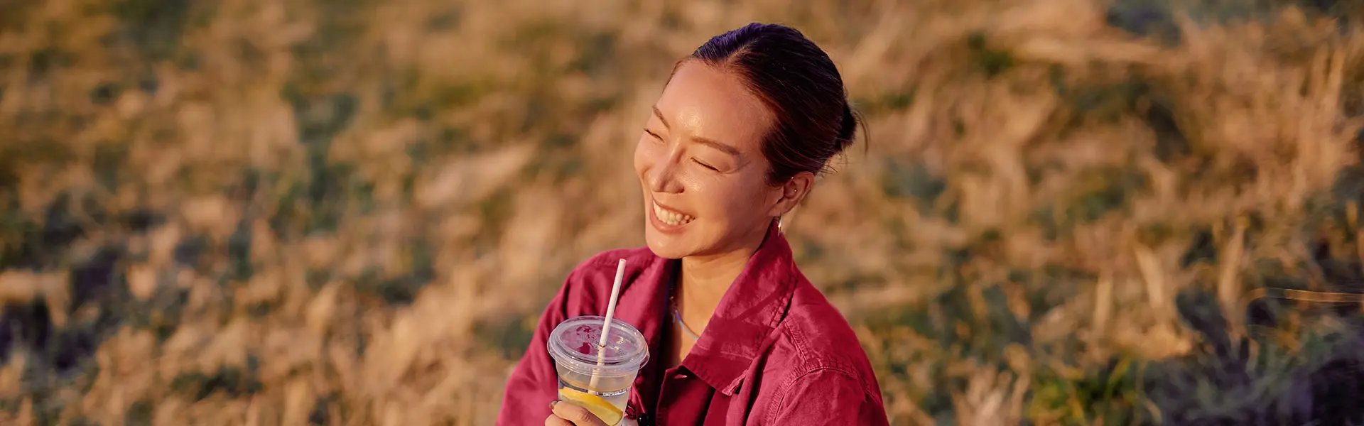 A person with a joyful expression holds a clear cup filled with a citrus drink, wearing a bright pink jacket. The background shows grass bathed in warm sunlight, creating a cheerful atmosphere.