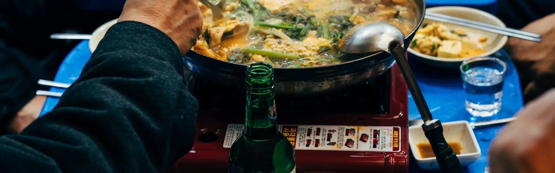 street-food scene with several people gathered around a small blue plastic table, cooking a large bubbling pot of stew on a portable gas burner; side dishes like kimchi and sauces surround the pot, along with bottles of soju, bowls, chopsticks, and napkins, capturing a busy, casual outdoor dining atmosphere.